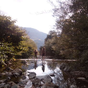 The image depicts two children standing in a shallow river, surrounded by rocks and lush greenery, with a mountainous landscape visible in the distance.