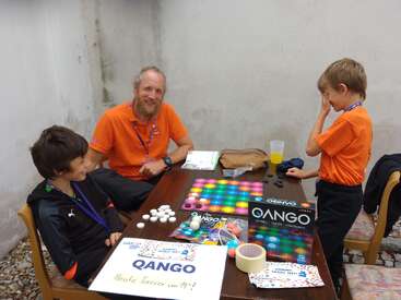 Three people, two children and one adult, are playing Qango board game at a table. They look engaged and happy. Promotional game materials are visible.