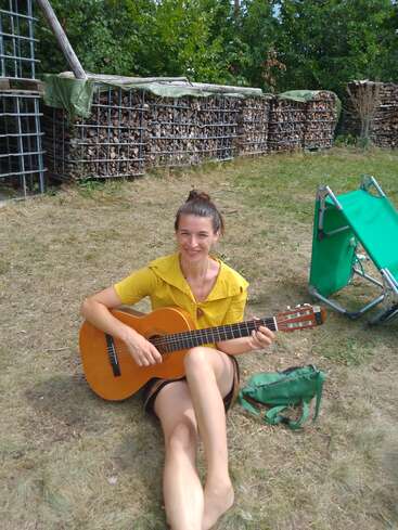 A woman sits barefoot on the grass, playing a guitar. Behind her are stacked logs and trees. She smiles, wearing a yellow shirt and holding a green bag.