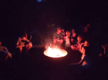 A group of people, mostly children, sit around a glowing campfire at night. They appear to be talking, relaxing, and roasting marshmallows together.