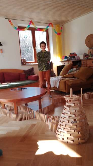 A boy stands in a sunlit, cozy living room. Wooden dominoes are arranged creatively on the floor, forming lines and a tower structure. Festive decorations hang above.