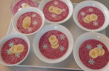 Seven bowls of pink smoothie or chia pudding, each topped with banana slices and white snowflake-shaped decorations, are arranged neatly on a white tray.