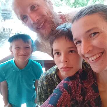 Ein glückliches Familien-Selfie mit zwei Erwachsenen und zwei Kindern. Alle lächeln, außer einem Kind, das ernst aussieht. Der Hintergrund scheint hell und im Freien zu sein.