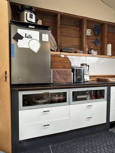 Cette cuisine est équipée d'un réfrigérateur orné d'aimants, d'un autocuiseur posé dessus, de tiroirs à façade en verre, d'une cafetière, d'étagères en bois, d'ustensiles de base et de touches rustiques en bois.