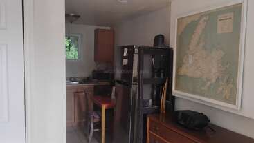 A small kitchen area with wooden cabinets, a chair, and a table is visible. A refrigerator stands nearby, with a large map and radio on a dresser.