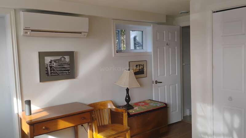 A cozy, sunlit room with a wooden desk, chair, table lamp, air conditioner, framed pictures, a small window, and an open closet and door.