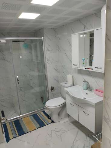Este é um banheiro moderno com azulejos com padrão de mármore, box de vidro, pia e vaso sanitário brancos, tapete listrado azul, armário espelhado e vários produtos de higiene pessoal. Iluminação clara.
