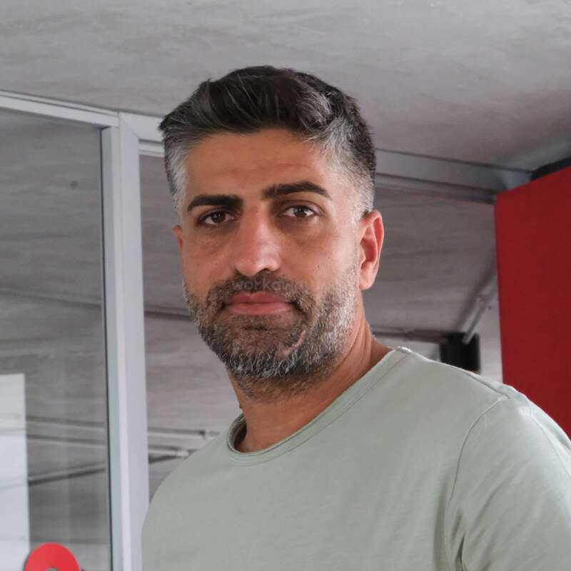 Um homem de cabelos curtos e escuros e barba está em um ambiente interno, vestindo uma camisa verde-clara. Ele encara a câmera com uma expressão neutra. O plano de fundo é moderno.
