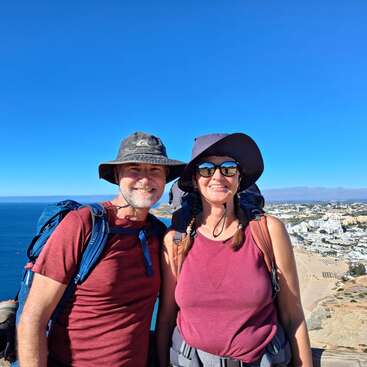 Un couple souriant, coiffé de chapeaux et équipé de sacs à dos, se tient debout en plein air, avec en arrière-plan un magnifique océan bleu et une ville balnéaire, sous un ciel clair et ensoleillé.