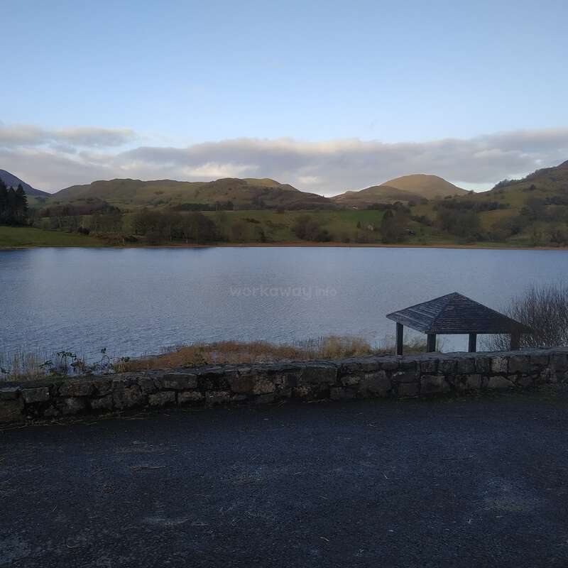 A imagem retrata uma cena serena de um lago com uma parede de pedra, um gazebo e colinas distantes sob um céu azul com nuvens, evocando uma atmosfera pacífica.