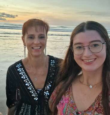 Duas mulheres estão sorrindo e tirando uma selfie em uma praia ao pôr do sol. As ondas do mar e o céu colorido criam uma atmosfera pacífica, alegre e calorosa.