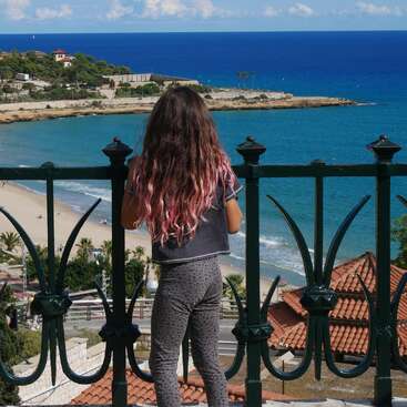 A girl with long hair partially dyed pink stands by a wrought iron railing, overlooking a scenic beach, blue sea, rooftops, and coastline under a sunny sky.