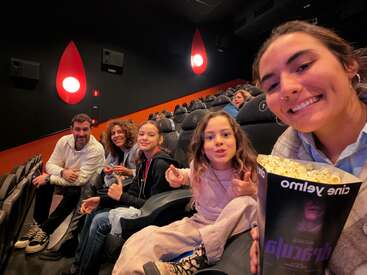 A group of five people, including children and adults, smile and pose for a selfie inside a movie theater, holding popcorn, ready to enjoy a film.