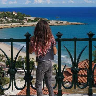 A girl with long hair partially dyed pink stands by a wrought iron railing, overlooking a scenic beach, blue sea, rooftops, and coastline under a sunny sky.