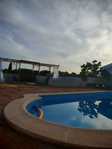 A tranquil backyard scene with a blue swimming pool, foam noodles, wooden pergola, sheer curtains, trees, cloudy sky, and serene reflections near sunset. Peaceful atmosphere prevails.
