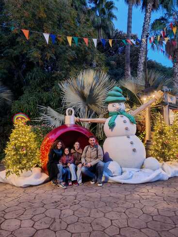 A family sits happily in front of a giant Christmas ornament, surrounded by festive trees, bright lights, a snowman decoration, and colorful flags under palm trees.