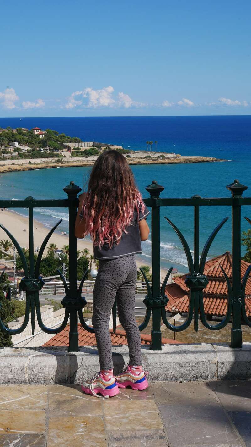 A girl with long hair partially dyed pink stands by a wrought iron railing, overlooking a scenic beach, blue sea, rooftops, and coastline under a sunny sky.