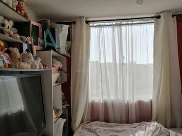 A imagem mostra um quarto com uma janela, cortinas brancas e prateleiras cheias de bichos de pelúcia, com uma cama com um edredom floral em primeiro plano.