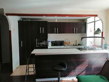 A imagem mostra uma cozinha moderna com armários de madeira escura, bancadas brancas e uma ilha central com banco de bar, pia e fogão.