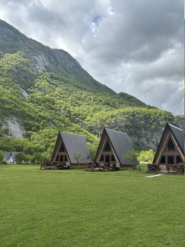 Three cozy A-frame cabins sit on a lush green field, surrounded by trees, with a backdrop of majestic mountains and a dramatic, cloudy sky above.