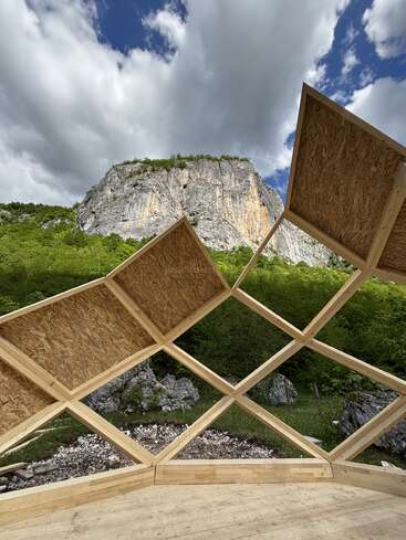 A wooden geometric framework stands in the foreground, partially sheltering a lush, green landscape. Behind, a tall rocky cliff rises under a dramatic cloudy sky.