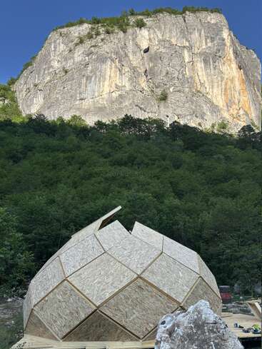 A partially constructed geometric wooden dome sits in a lush, green forest at the base of a massive rocky cliff, beneath a clear blue sky.