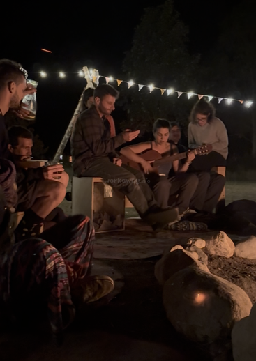 A group of friends sit around a campfire at night, playing guitar, drinking, and enjoying music together, surrounded by string lights and festive flags.