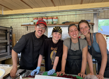 Four smiling people pose inside a kitchen, wearing aprons and gloves, preparing food together. Shelves, canned tomatoes, and ovens are visible in the cozy background.