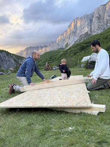 Two men and a small child are building with wooden boards outdoors, surrounded by beautiful mountains, green grass, and triangular wooden cabins under a dramatic sky.