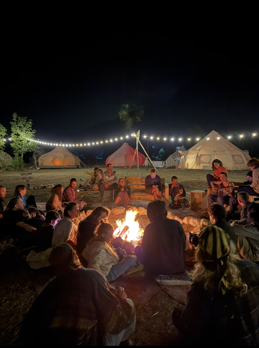A group of people sit around a glowing campfire at night, surrounded by tents and string lights, enjoying music, warmth, and conversation in a cozy campsite.