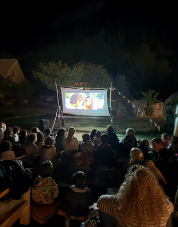 A group of people gathers outdoors at night, watching an animated movie projected on a screen, surrounded by nature and cozy atmosphere, creating a communal experience.