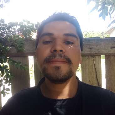 A man with dark hair and facial hair stands outside in front of a wooden fence, surrounded by leafy green branches, sunlight casting shadows on his face.