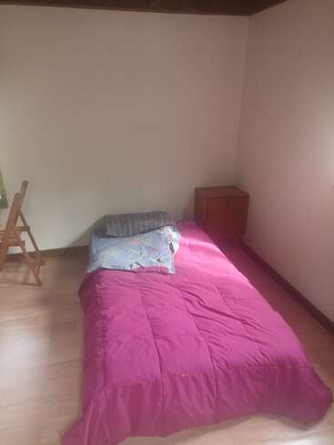This image shows a simple bedroom with a single bed covered by a pink blanket, a small nightstand, a wooden chair, and plain white walls.