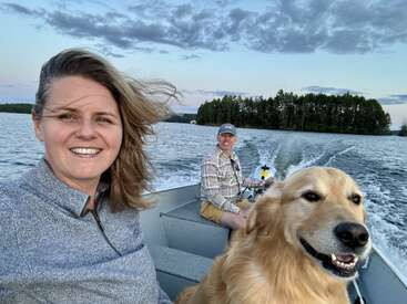 Eine Frau, ein Mann und ein Golden Retriever machen eine Bootsfahrt auf einem See. Der Himmel ist bewölkt, und im Hintergrund sieht man Bäume.