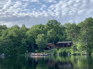 Eine ruhige Szene am See zeigt eine gemütliche Holzhütte, die zwischen üppig grünen Bäumen liegt, einen Steg mit Booten und ruhiges Wasser, das die friedliche Natur umgibt.