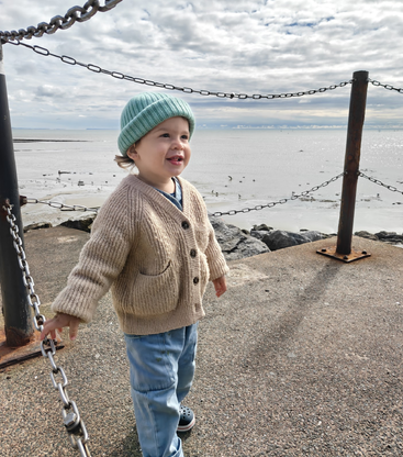A young child in a beige sweater and blue hat stands near the water, smiling, surrounded by chains and rocks under a cloudy, bright sky.