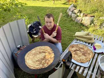 A person sits outdoors with crossed spatulas, cooking large pancakes on a griddle. Nearby, baked pancakes, butter, and oven mitts rest on a wooden table.