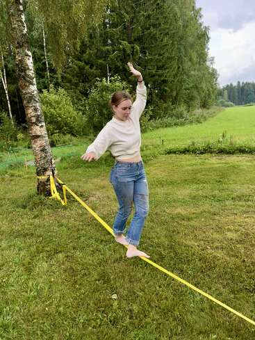 A woman balances barefoot on a yellow slackline tied between two trees, with green grass and a forest in the background on a cloudy day.