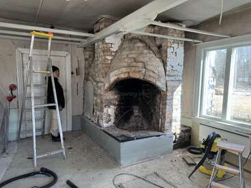 A person stands in a room under renovation, next to a tall metal ladder and a large, old, partially exposed brick fireplace, with sunlight streaming in.