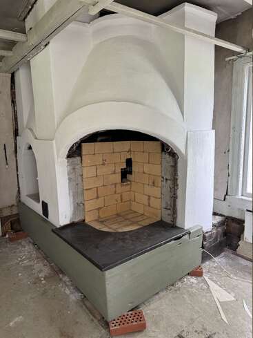 This image shows a traditional, white-painted masonry stove with a brick firebox and a green wooden base, located in a partially unfinished room with exposed areas.