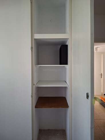 This image shows an empty built-in white closet with four shelves, one shelf containing a black object. The closet is in a plain, minimally decorated room.