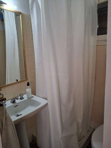 This image shows a small bathroom with a pedestal sink, a wall mirror, beige tiles, a white shower curtain, a towel, soap dispenser, and toilet.