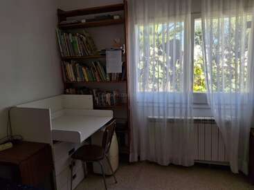 A cozy study corner with a white desk, wooden chair, bookshelf filled with books, and bright window with sheer curtains letting in natural daylight. Peaceful atmosphere.