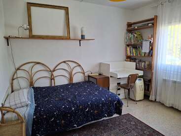 This cozy bedroom features a rattan bed with a starry night comforter, a desk, bookshelves filled with books, a window with sheer curtains, and minimalist decor.