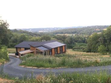 A imagem mostra uma casa moderna com painéis solares no telhado, situada em uma área rural cercada por árvores e uma entrada de automóveis sinuosa.