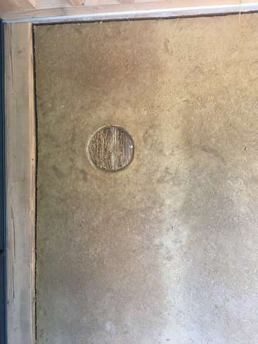 A imagem mostra uma parede com um recorte circular, com um material marrom claro que lembra concreto ou gesso, cercado por molduras de madeira e um teto de madeira.