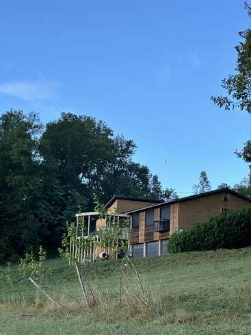 A rustic house sits on a grassy hill, surrounded by trees under a clear blue sky. A small balloon floats high in the distance.