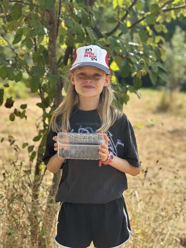 A young girl stands outdoors, wearing a cap and dark clothes, holding a transparent container. She is surrounded by greenery, with sunlight filtering through the trees.