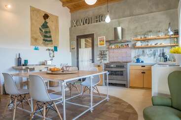 Cette cuisine moderne et chaleureuse présente des accents en bois et en métal, des étagères ouvertes, une grande table en bois, des chaises minimalistes, des œuvres d'art murales et une lumière naturelle intense dans toute la pièce.