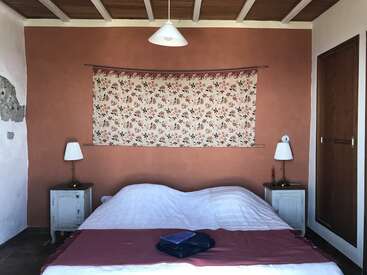 Se trata de un acogedor dormitorio con cama de matrimonio, dos mesillas de noche, lámparas blancas, colgantes florales, toallas dobladas, pared rústica naranja y techo de madera.